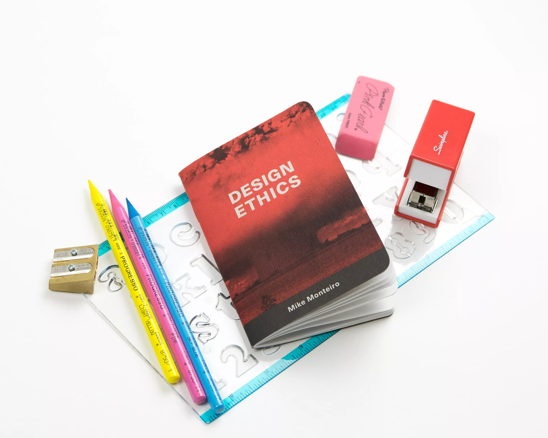 A red booklet titled “Design Ethics” by Mike Monteiro rests on a desk surrounded by colourful pencils, a ruler, sharpener, and stapler.