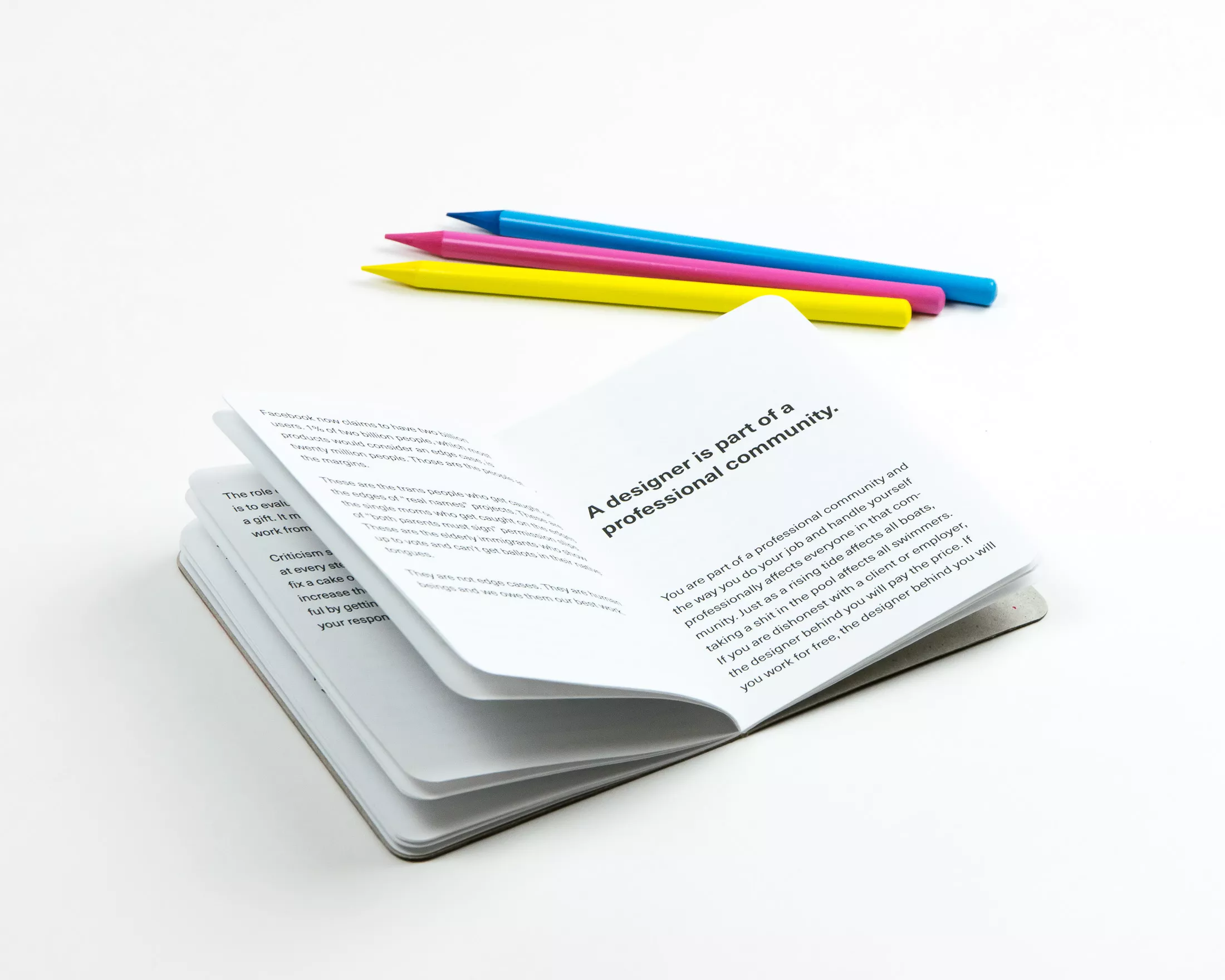Open booklet showing the page “A designer is part of a professional community” alongside brightly coloured pencils on a white surface.